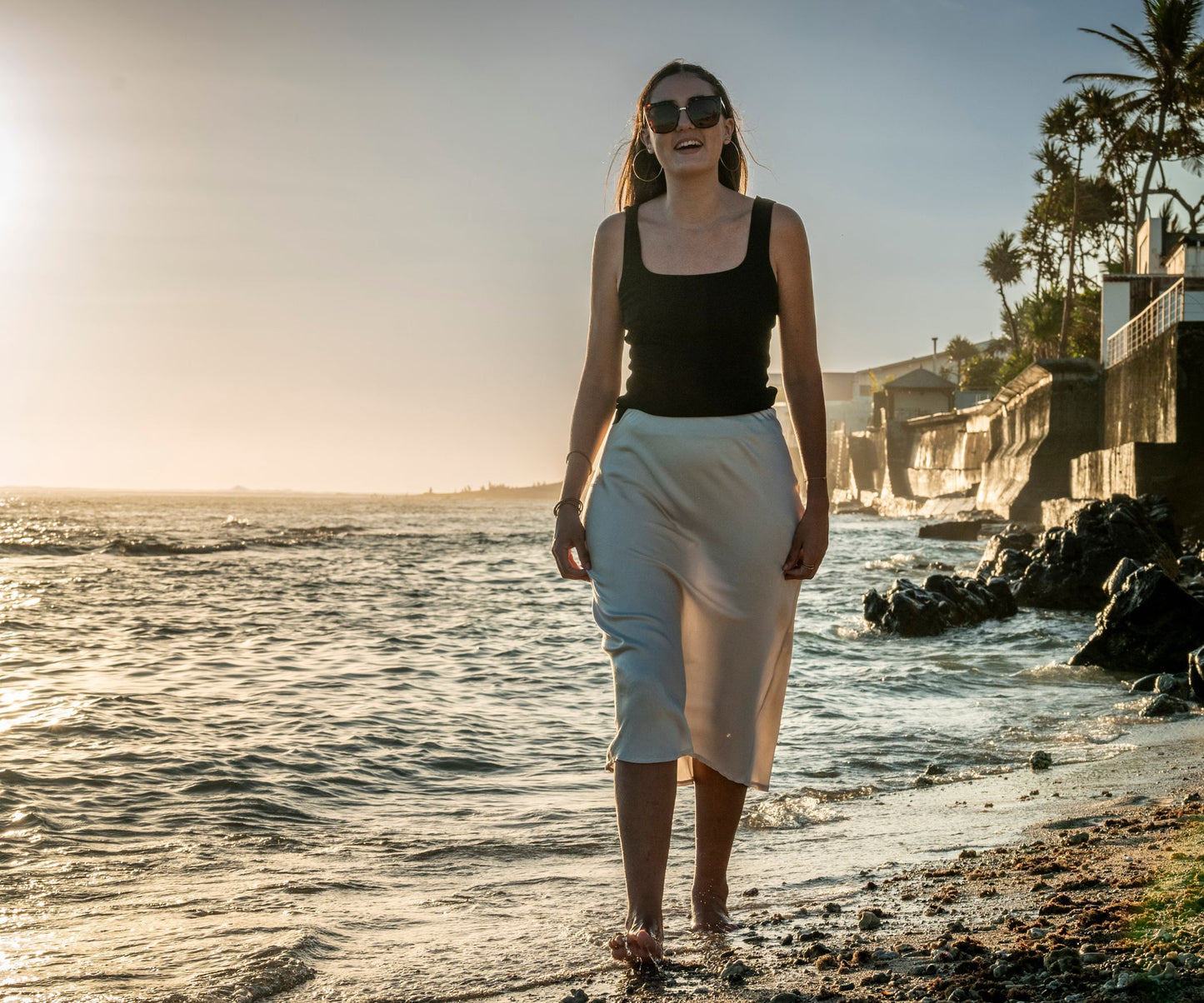 Femme sur la plage les pieds dans l'eau portant une jupe effet soie beige et un top noir