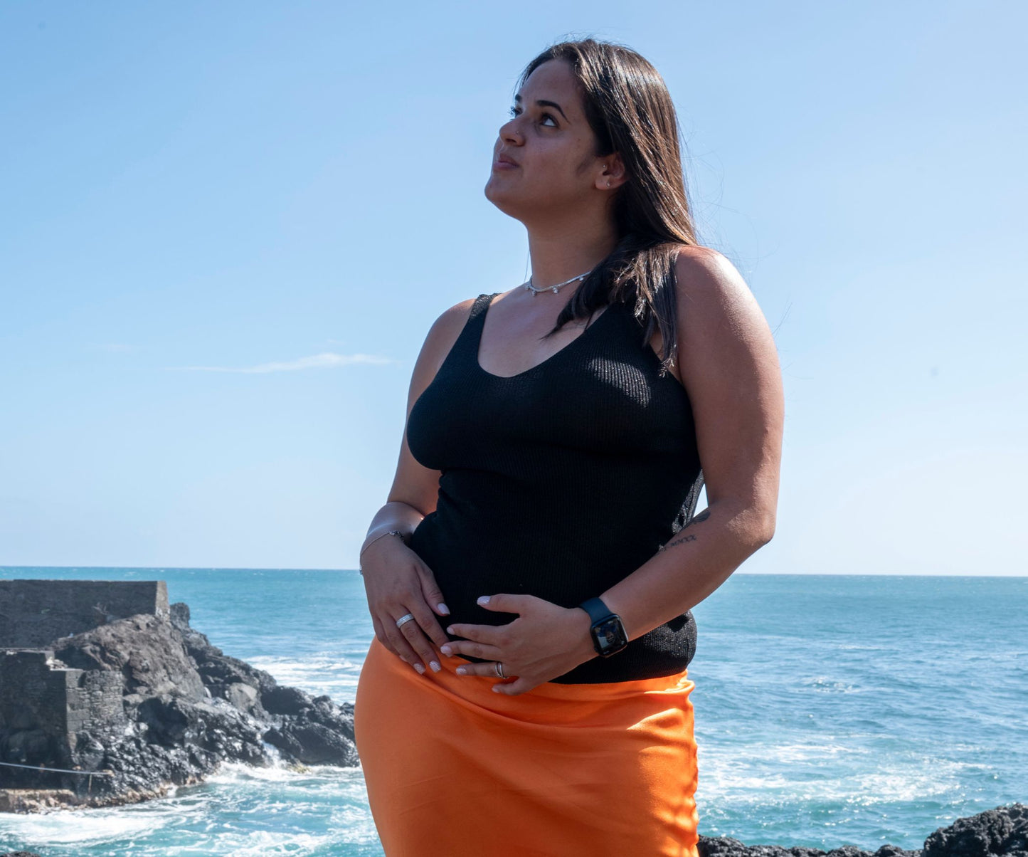 Femme portant un top col V noir avec fil lurex et jupe orange effet soie avec l'océan en arrière plan